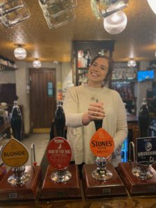 The British Oak's General Manager, Kate, standing behind the bar, pulling a cask pint of Stones.